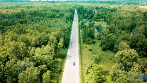 Wide-shot-aerial-drone-footage-tracking-a-sedan-car-cruising-on-an-empty-road-surrounded-by-a-beautiful-large-forest-and-river
