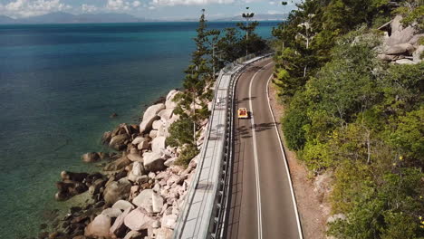 Aerial-view-of-open-top-convertible-classic-beach-buggy-driving-along-coastal-clifftop-road-next-to-crystal-clear-tropical-water-and-lush-green-forest