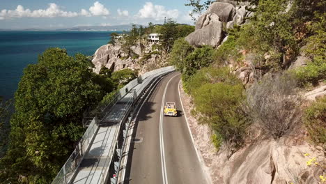 Aerial-view-of-young-woman-driving-open-top-convertible-classic-beach-buggy-along-coastal-clifftop-road-next-to-crystal-clear-tropical-water