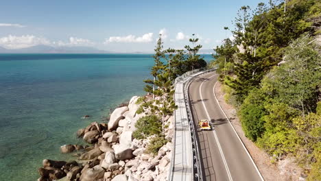 Aerial-view-of-open-top-convertible-classic-beach-buggy-driving-along-coastal-clifftop-road-next-to-crystal-clear-tropical-water-with-bright-blue-sky