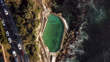 AERIAL:-Drone-footage-of-Bronte-Sea-Baths-with-swimmers-showing-contrast-between-wild-ocean-and-urban-development