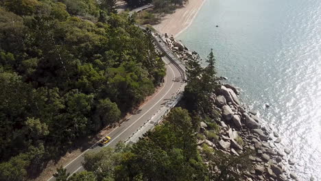 Aerial-view-of-open-top-convertible-classic-beach-buggy-driving-along-coastal-clifftop-road-next-to-with-sun-reflecting-off-crystal-clear-tropical-water