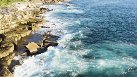 While-fringed-waves-crashing-onto-ocean-rocks-POV-drone-moving-slowly-backwards-along-cliff-face-ocean-pool-in-background-Coogee-Beach-Sydney-Australia
