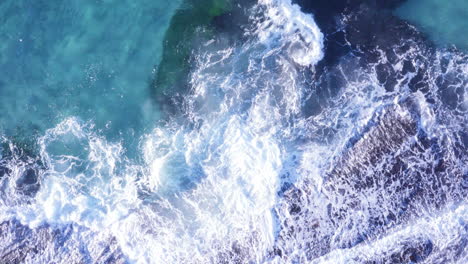 White-tipped-waves-roll-onto-dark-ocean-rocks-clear-blue-water-Tamarama-beach-Sydney-Australia-POV-drone
