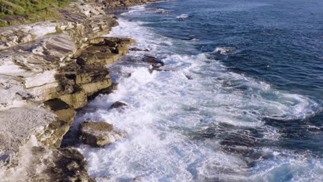 While-fringed-waves-crashing-onto-ocean-rocks-POV-drone-moving-slowly-forward-along-cliff-face-ocean-pool-in-background-Coogee-Beach-Sydney-Australia