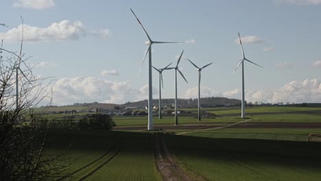 Windkraftanlagen-Drehen-Sich-Auf-Einem-Feld-Mit-Weg-Zum-Dorf-Im-Hintergrund-Und-Bäumen-Im-Vordergrund