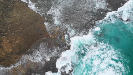 A-sweeping-aerial-top-view-of-waves-crashing-against-a-rock-face-on-a-tropical-island