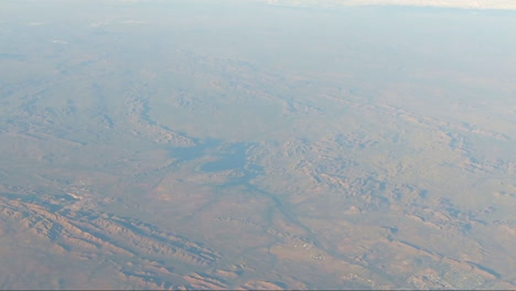 POV-from-aircraft-window-showing-red-earth-mountain-range-and-lake-mid-screen-Town-bottom-right-of-screen-with-river-flowing-into-town-from-mid-screen-outback-Australia