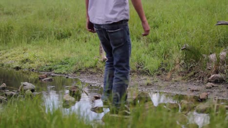 Young-man-crosses-in-front-of-camera,-traverses-stream,-and-walks-away-into-field