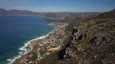 Drone-descending-behind-Silver-Mine-Mountains-with-coastal-suburb-St-James-behind,-Cape-town