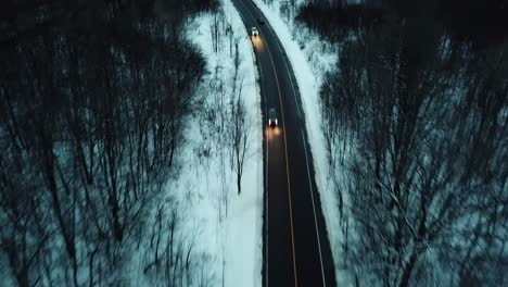 Birdseye-view-drone-shot-of-cars-passing-on-secluded-road-way-in-winter-forest-in-the-dark