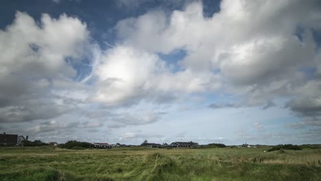 Timelapse-of-clouds-moving-towards-the-viewer-in-a-blue-sky-over-houses-with-gras-in-foregorund