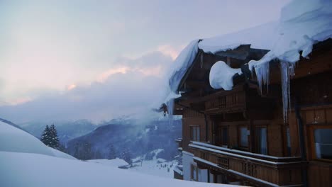 Snow-covered-wooden-chalet-with-icicles-and-snow-blown-by-the-wind-during-sunset