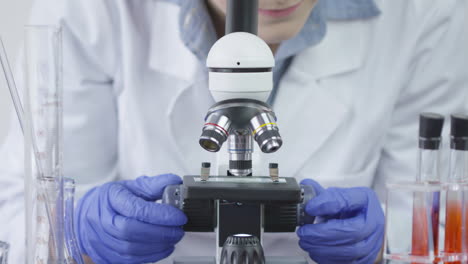 Ein-Labortechniker-Mit-Blauen-Handschuhen-Dreht-Langsam-An-Den-Knöpfen-Eines-Mikroskops-Mit-Reagenzgläsern-Daneben