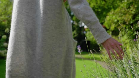 Slow-motion-close-up-shot-following-a-young-woman-walking-through-a-lush-green-garden,-moving-her-hand-through-the-plants-vegetation-high-grass