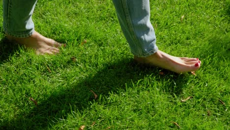 Slow-motion-close-up-side-shot-following-a-woman-walking-barefoot-in-jeans-through-a-green,-lush-lawn-casting-a-shadow-towards-the-camera