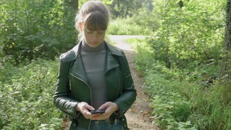 Young-woman-stares-down-at-smartphone-in-hand-in-forest,-tracking,-medium