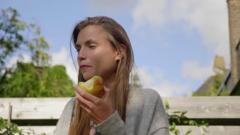 Young-woman-eats-apple-sitting-outside-enjoying-fresh-breeze,-pan