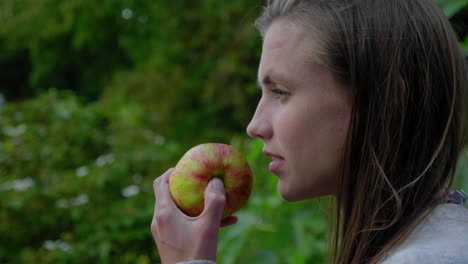 Young-womans-bites-apple-and-stares-thoughtfully-in-garden,-profile