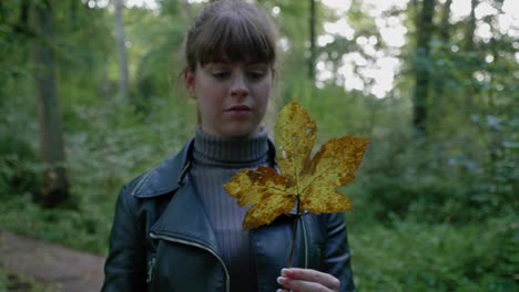 Beautiful-young-woman-twirls-gold-sweatgum-leaf-in-forest,-tracking