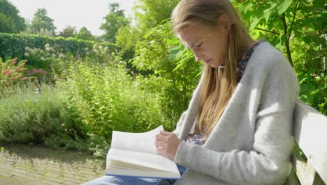 Slow-motion-medium-shot,-from-the-side,-of-an-attractive-young-woman-reading-and-studying-in-a-lush-green-garden-environment-turning-pages