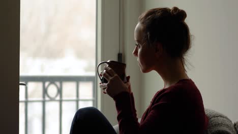 Medium-shot-of-young-woman-quietly-watching-snowflakes-fall-outside-and-enjoying-hot-beverage-in-mug