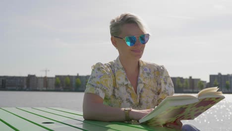 Young-millennial-woman-enjoys-a-novel-while-sitting-next-to-a-river