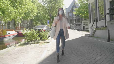 Woman-Wearing-Mask-Walking-Alone-At-The-Promenade-Of-Amsterdam-Canal---Coronavirus-Pandemic-In-Netherlands---full-shot,-slow-motion