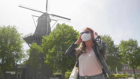 Young-woman-puts-on-face-mask-with-Dutch-windmill-behind,-low-angle