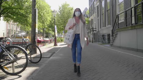 Erwachsene-Blonde-Dame-Mit-Gesichtsmaske-Geht-Während-Des-Covid19-ausbruchs-Auf-Ziegelpflaster-In-Amsterdam,-Niederlande