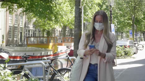 Schöne-Dame-Mit-Medizinischer-Maske,-Die-Inmitten-Einer-Covid-19-pandemie-Die-Straße-In-Der-Nähe-Des-Amsterdamer-Kanals-In-Den-Niederlanden-Entlang-Geht