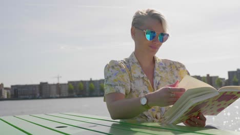 Stylish-young-blonde-woman-reads-a-book,-sitting-at-a-picnic-table,-river-in-the-background