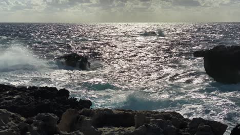 Sunlight-reflects-off-blue-ocean-crashing-against-backlit-rocky-coast,-Gozo-Malta