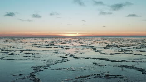 Drone-flying-over-calm-sea-with-melted-ice-at-the-surface-at-sunset,-sunrise