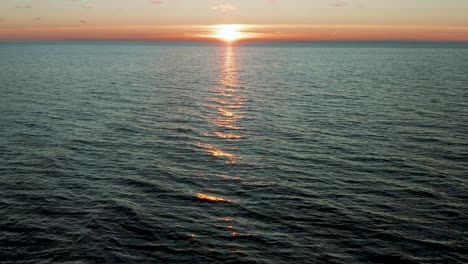 Drone-flying-over-calm-wavy-sea,-camera-tilt-up-to-a-beautiful-sunset,-sunrise