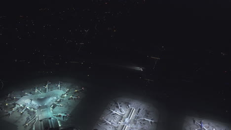 Eine-Vogelperspektive-Eines-Flughafens-Mit-Mehreren-Terminals-Mit-Geparkten-Flugzeugen-Und-Einem-Flugzeug,-Das-Sich-Nachts-Bewegt