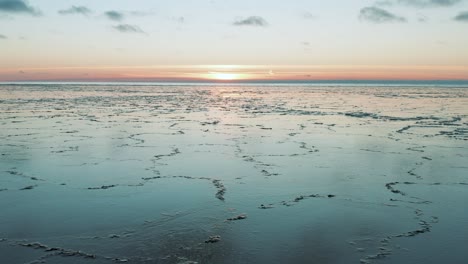 Drone-flying-over-calm-sea-with-melted-ice-at-the-surface-at-sunset,-sunrise