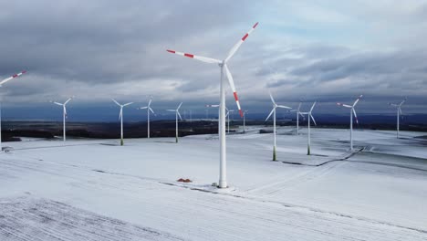 Aerial-backwards-shot-of-rotating-wind-turbine-farm-in-winter-with-snow-covered-and-frozen-agricultural-fields
