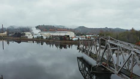 Aerial-orbit-of-old-swing-bridge-in-the-middle-of-foggy-lake-near-small-factory