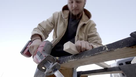 Male-carpenter-cuts-through-wood-with-reciprocating-saw-outside,-low-angle-shot