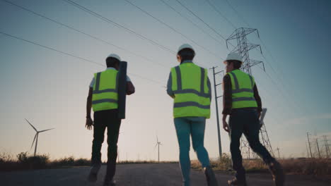 Supervisors-wearing-white-hardhats-and-green-vests-walking-down-a-gravel-path-under-transmission-towers-next-to-wind-turbines-at-sunset