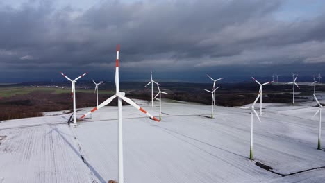 Aerial-high-angle-circling-shot-of-spinning-wind-turbines-working-on-snow-covered-rural-fields-during-sunny-day-with-clouds-in-winter