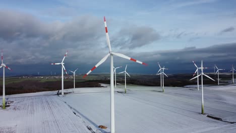 Aerial-view-of-rotating-wind-turbines-standing-on-iced-and-snowcapped-rural-field-in-nature