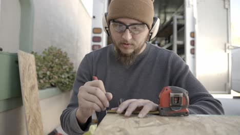 Craftsman-wearing-safety-glasses-and-ear-muffs-marks-timber-with-pen-and-tape-measure