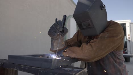 Man-wearing-welders-jacket,-gloves,-and-hood-welds-angle-iron-together-outside,-slow-mo