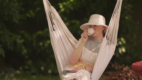 A-middle-aged-casually-looking-woman,-wearing-a-striped-dress-and-a-sun-hat,-sitting-in-the-hammock-chair,-drinking-coffee-from-a-small-white-cup
