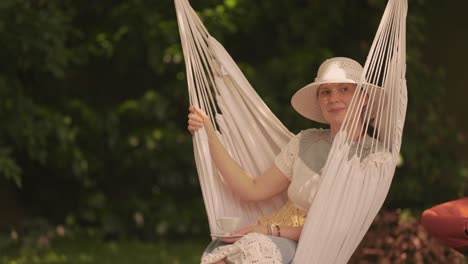 A-middle-aged-casually-looking-woman,-wearing-a-knitted-dress-and-a-sun-hat,-sitting-in-the-hammock-chair