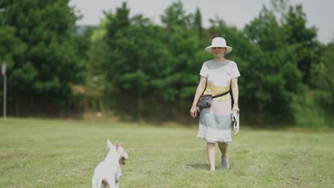 A-middle-aged-casually-looking-woman,-wearing-a-handmade-knitted-dress-and-a-wide-brimmed-sun-hat,-enjoying-the-walk-in-the-town-park-with-her-dog