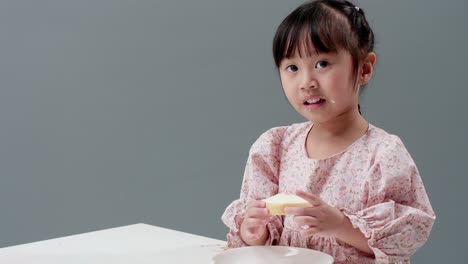 Asiatisches-Kind,-Das-Im-Studio-Ein-Stück-Süßes-Mit-Grauem-Hintergrund-Isst