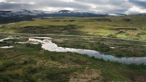 A-glimpse-on-the-tranquil-nature-of-the-Arctic-circle:-a-shallow-river-is-calmly-running-near-a-railway,-an-asphalt-road-is-neighboring-with-green-hillside,-snowy-peaks-are-touching-cloudy-sky
